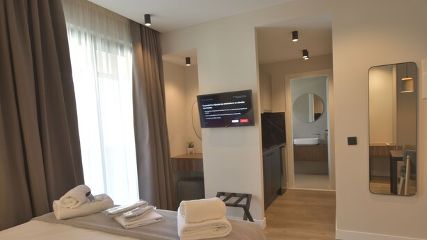 A modern hotel room with a neatly made bed, a flat-screen TV mounted on the wall, a small table with towels, and a view of the bathroom through an open door. - Charming Studio with Balcony and Acropolis View (Athens)