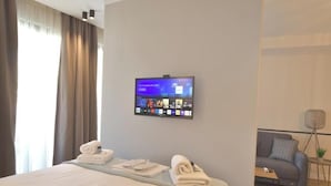 Ein Hotelzimmer mit Bett, einem an der Wand befestigten Flachbildfernseher, der eine Streaming-Plattform anzeigt, einer Couch und einem kleinen Tisch mit Lampe.