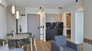 A modern hotel room with a round dining table, two armchairs, a sofa, a kitchen area with dark cabinets, and a ceiling-mounted air conditioning unit. - Superior Apartment with City View (Athens)