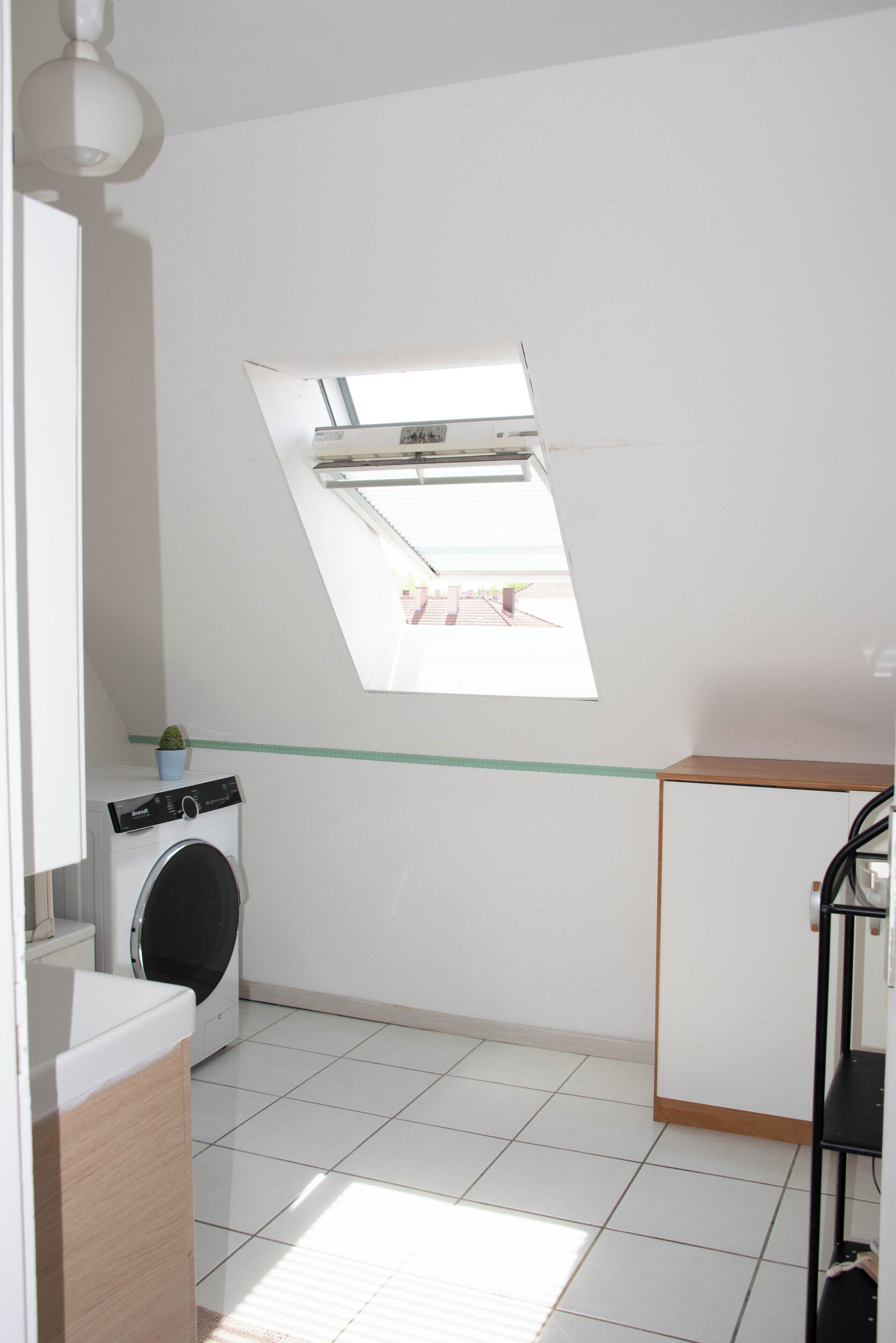 Een wasserette met een wasmachine, een kleine potplant, een dakraam en een betegelde vloer.