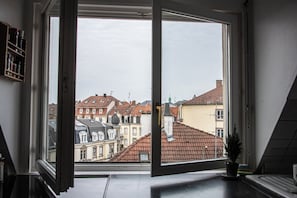 A room with a large window, a potted plant on the windowsill, and a view of neighboring buildings.
