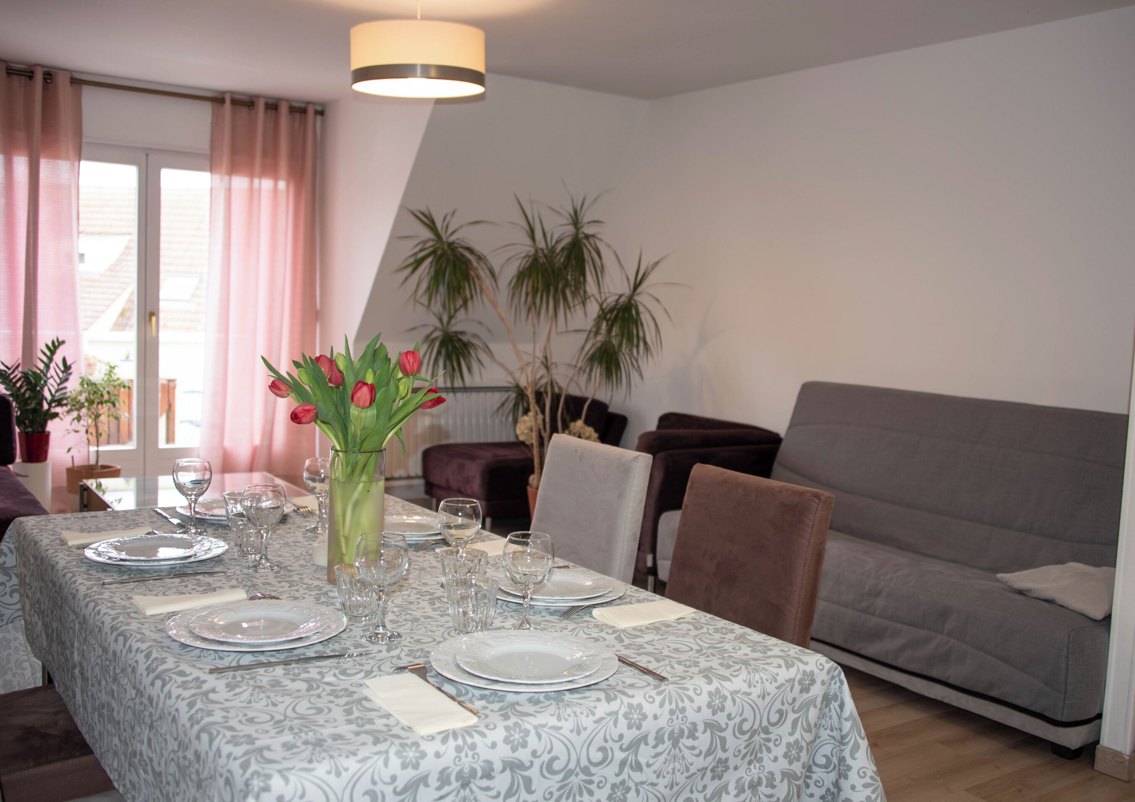 Uma sala de jantar com mesa posta para seis pessoas, um vaso com flores e um sofá cinzento ao fundo.