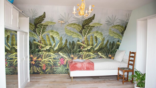 Un dormitorio con una cama, una silla de madera, una planta en maceta, un candelabro y un mural con follaje tropical en la pared.