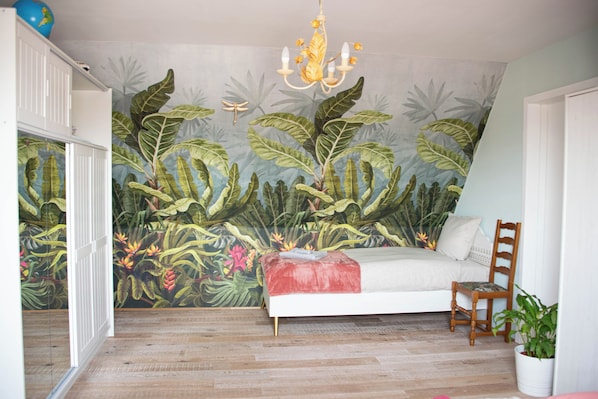 A bedroom with a bed, a wooden chair, a potted plant, a chandelier, and a mural of tropical foliage on the wall.
