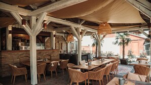 Un espace de restauration en plein air avec des tables en bois et des chaises en osier, un mur en briques et une grande fenêtre donnant sur un jardin.