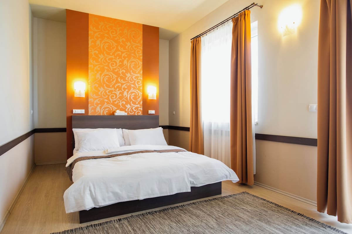 A hotel room with a large bed, two wall-mounted lights, a patterned wall, and a window with curtains.