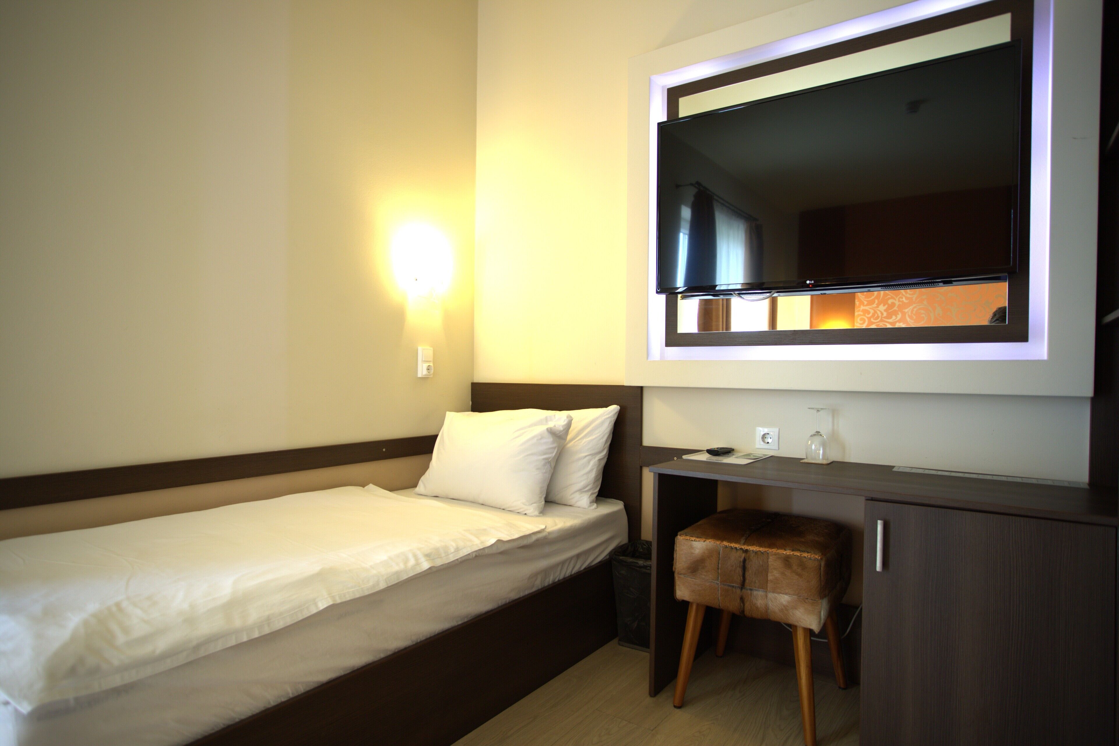 A hotel room with a single bed, a desk with a chair, a large mirror, and a wall-mounted TV.