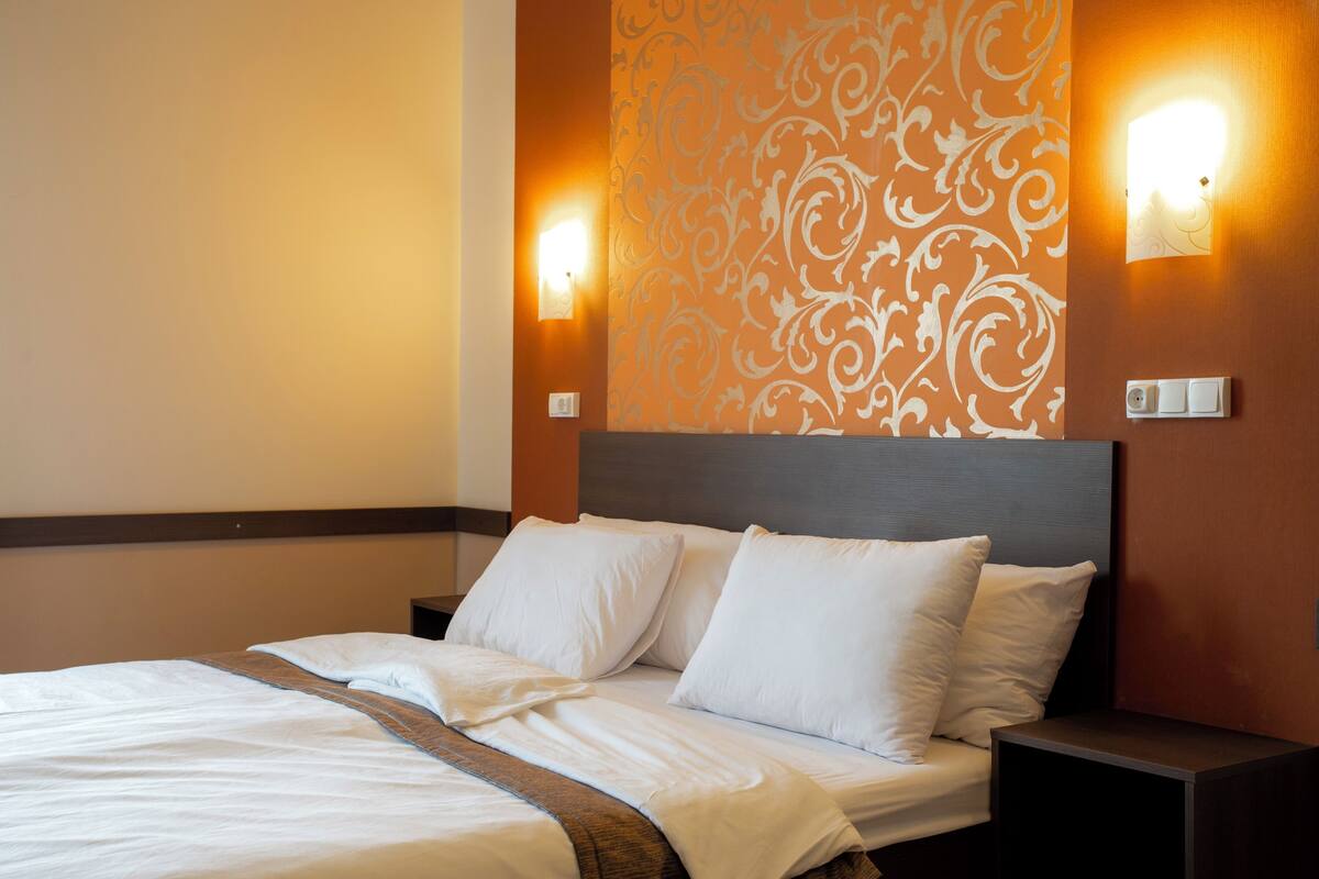 A neatly made bed with white linens and pillows, a wooden headboard, and two wall-mounted nightstands with light switches.