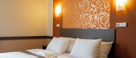 Una cama bien hecha con sábanas y almohadas blancas, un cabecero de madera y dos mesitas de noche fijadas a la pared con interruptores de luz.