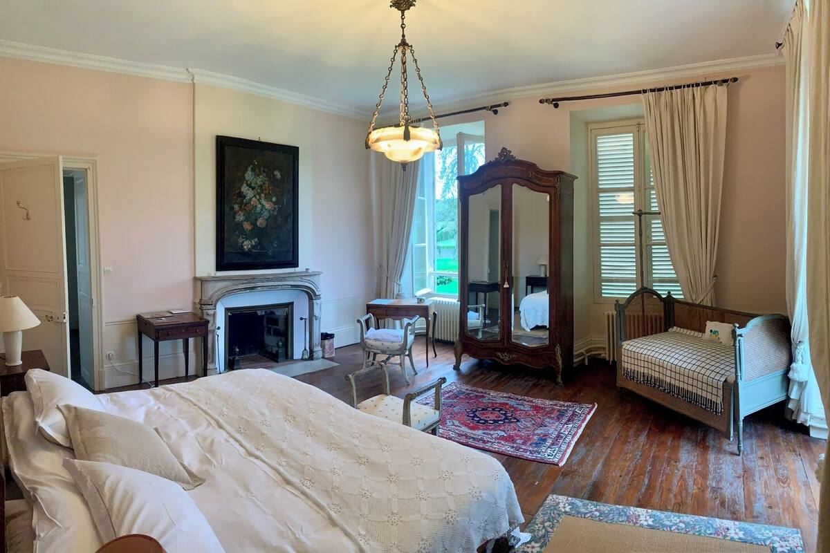 A traditional bedroom with a bed, a chair, a small table, a mirror, and a fireplace.