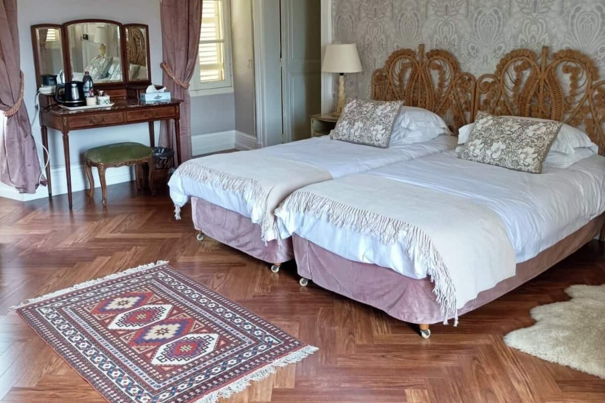 A bedroom with a double bed, a wooden desk with a mirror, a chandelier, and patterned wallpaper.