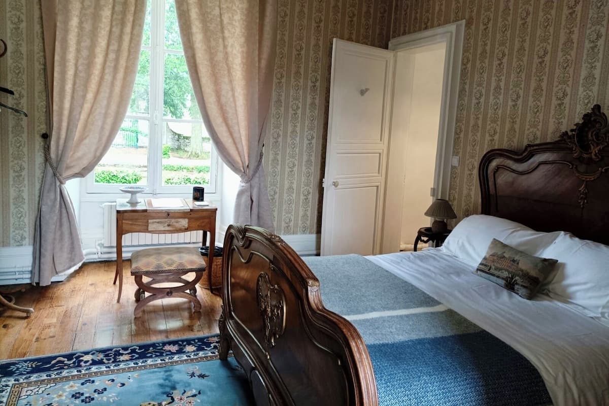 A bedroom with a wooden bed, a desk, and a chair. There is a window with curtains, a door, and a patterned wallpaper.