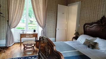 A bedroom with a wooden bed, a desk, and a chair. There is a window with curtains, a door, and a patterned wallpaper.