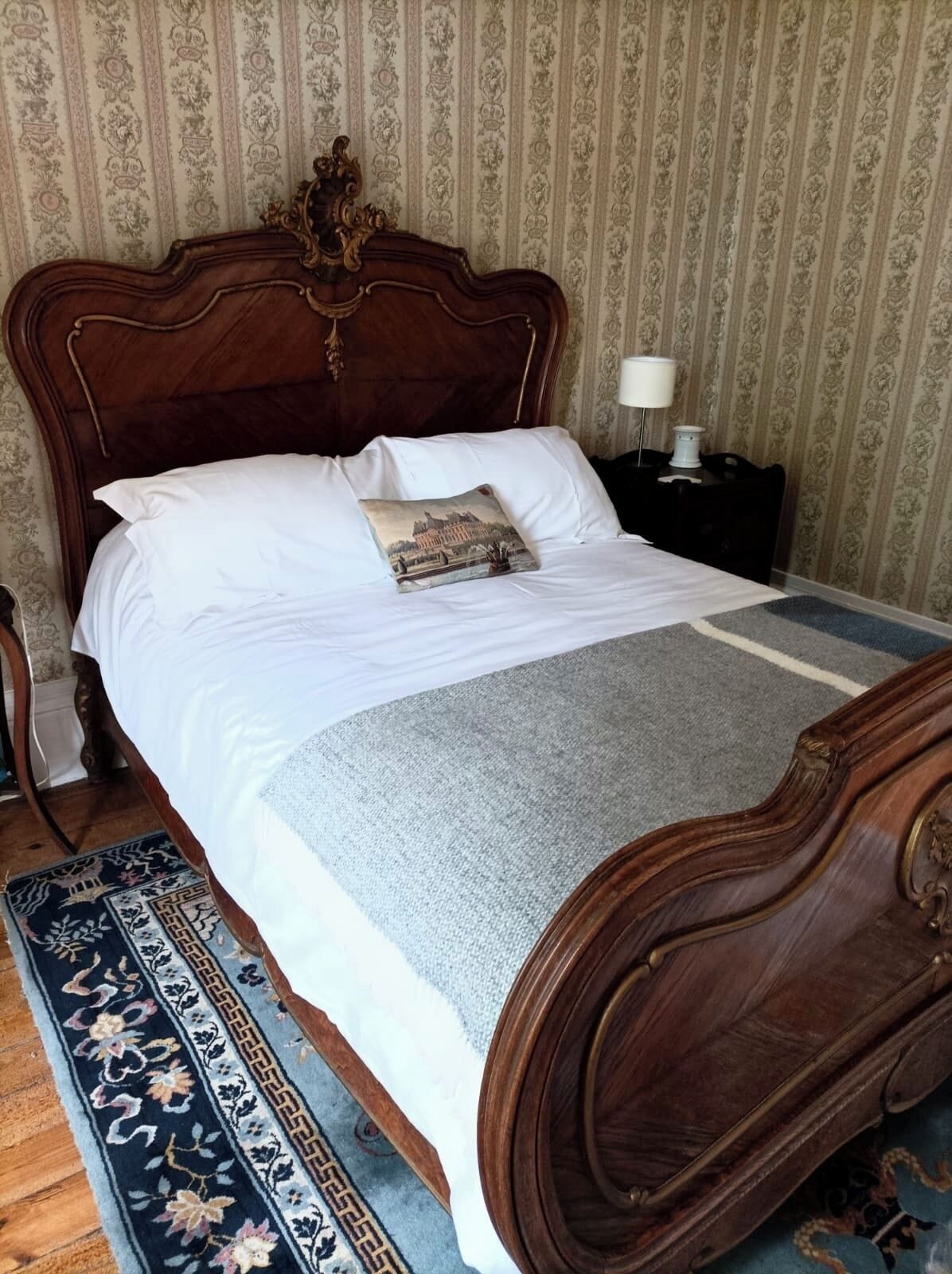 Un lit avec une tête de lit en bois, une literie blanche, un coussin décoratif, une couverture grise et un tapis à motifs.