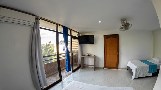 A hotel room with two beds, a TV mounted on the wall, a small table, and a balcony with a view of the outside. - Hotel Santander Plaza (Neiva)