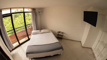 A bedroom with a bed, a TV, a balcony with a view of greenery, and a tiled floor.