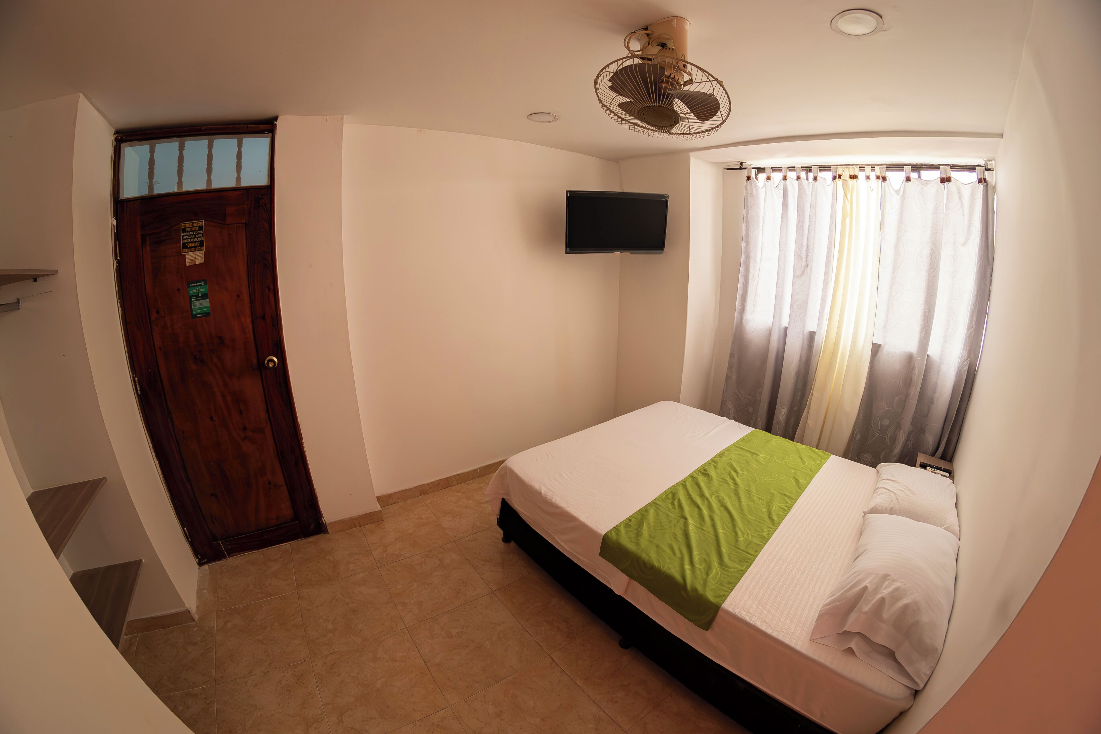 A bedroom with a bed, a television, a ceiling fan, and a window with curtains.