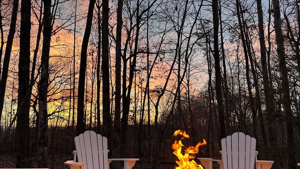 Ein Lagerfeuer in einer runden Feuerstelle aus Stein, mit zwei weißen Adirondack-Stühlen davor, umgeben von einem Wald.