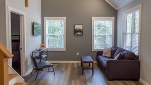 Ein Wohnzimmer mit einem dunkelbraunen Sofa, einem kleinen Tisch und einem Sessel. Zwei Fenster mit Jalousien und ein Bilderrahmen an der Wand sind zu sehen.