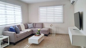 Ein modernes Wohnzimmer mit einem grauen Ecksofa, einem weißen Couchtisch und einem Flachbildfernseher auf einem weißen Schrank.