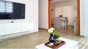 Ein modernes Wohnzimmer mit einem Flachbildfernseher an einem weißen Schrank, einem hölzernen Bogen, der in einen Essbereich führt, und einem Couchtisch mit einer Vase und einer Pflanze.