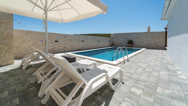A pool area with white lounge chairs and an umbrella, stone walls, and a clear sky.