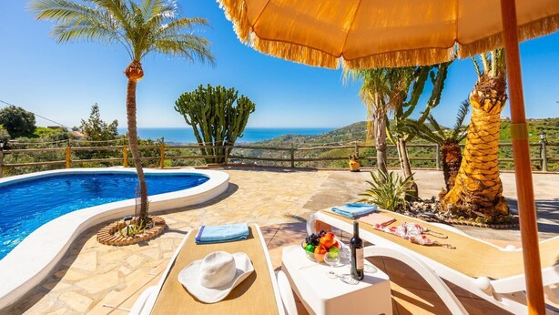 Ein Poolbereich mit Meerblick, einem Reetdachschirm und Liegestühlen.