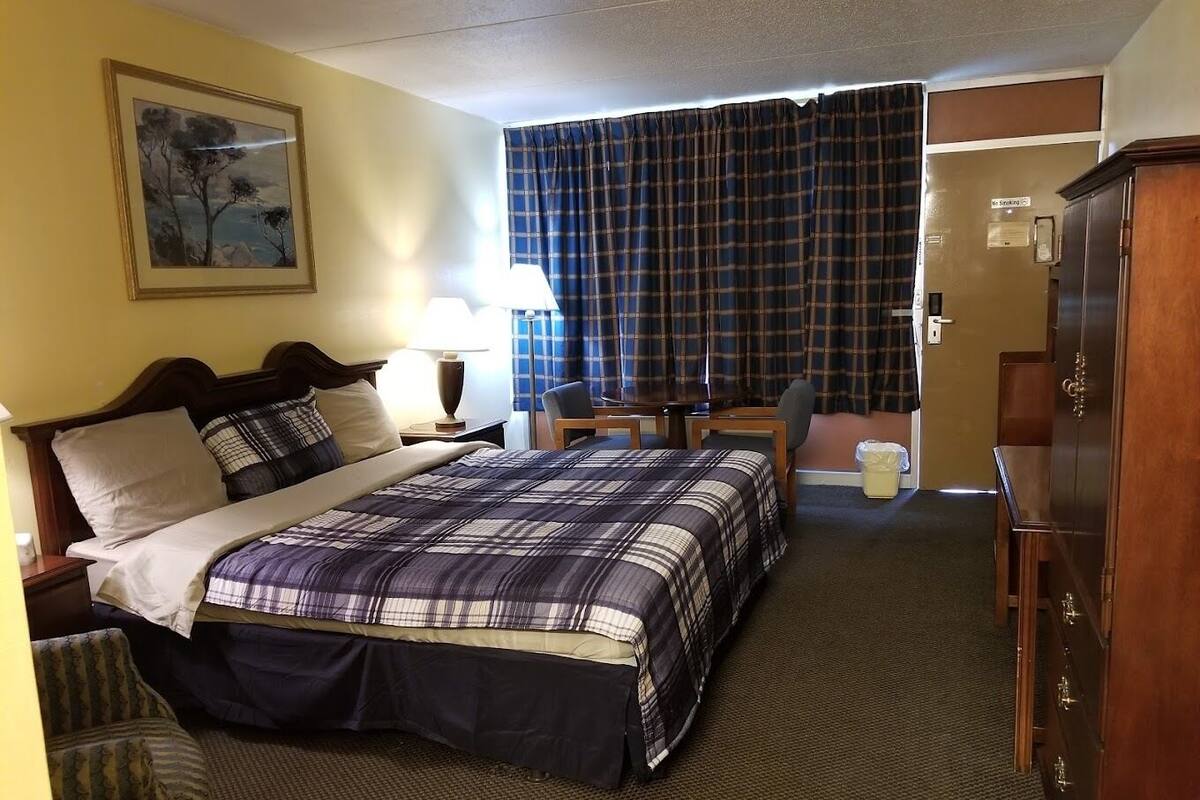 A hotel room with a bed, a desk, a chair, a wardrobe, a lamp, and a painting on the wall.
