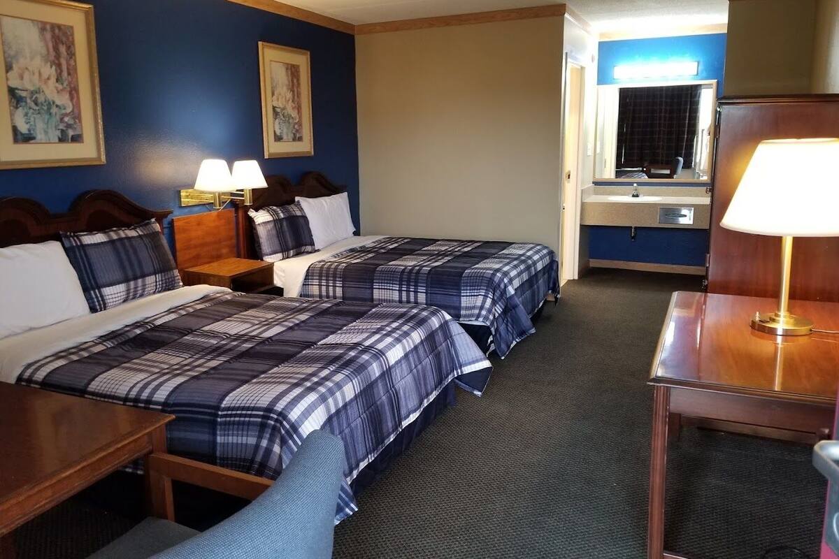 A hotel room with two beds, a desk, a chair, a nightstand, a lamp, a mirror, and a bathroom visible through an open door.