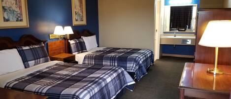 A hotel room with two beds, a desk, a chair, a nightstand, a lamp, a mirror, and a bathroom visible through an open door.