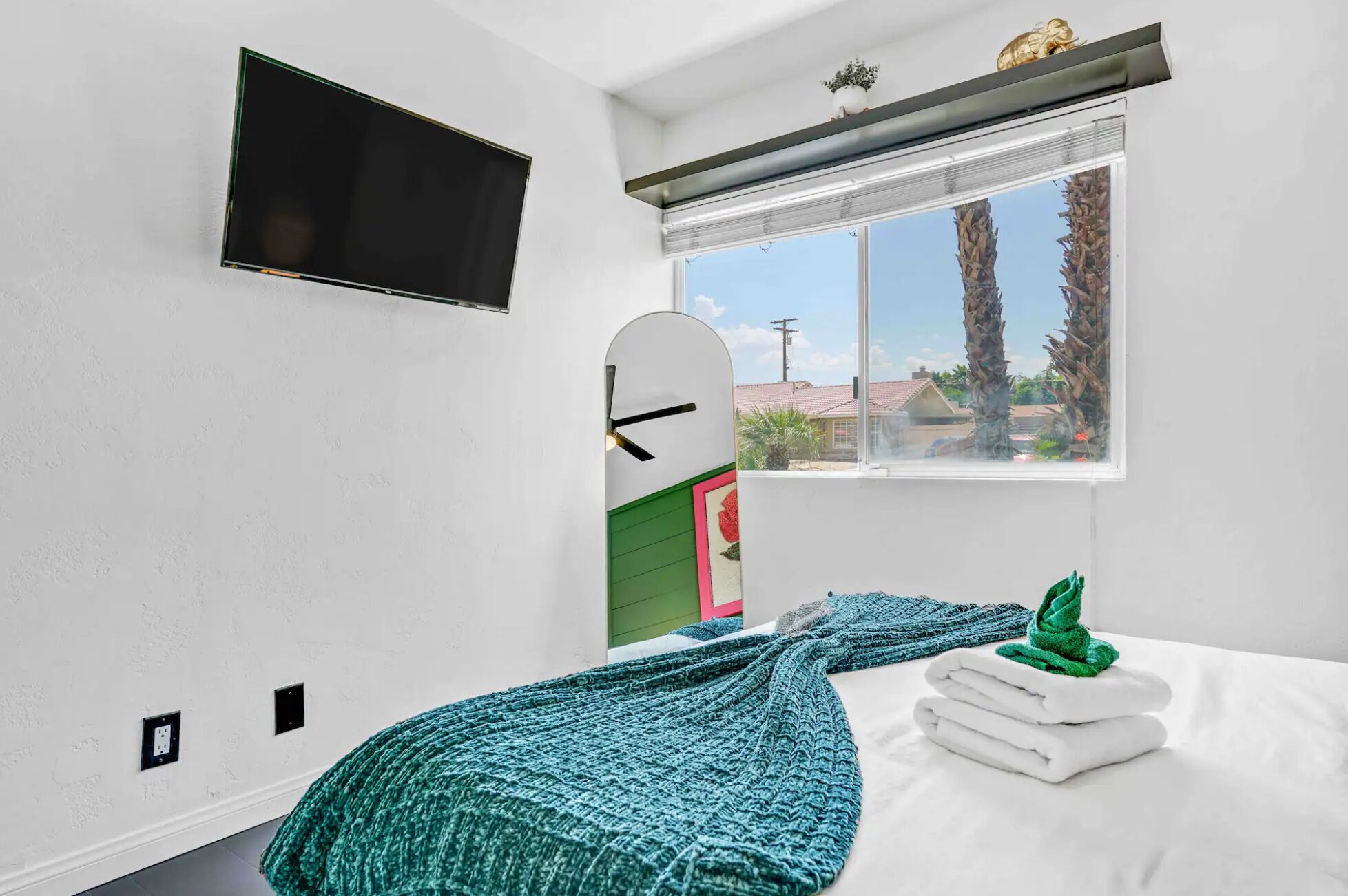 A bedroom with a bed, a window with a view, a mounted TV, and a green knitted blanket.