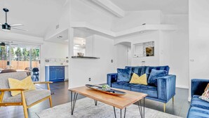 Amplia sala de estar con un sofá azul, una mesa de centro de madera y un sillón gris. Se ve una cocina al fondo con armarios azules.