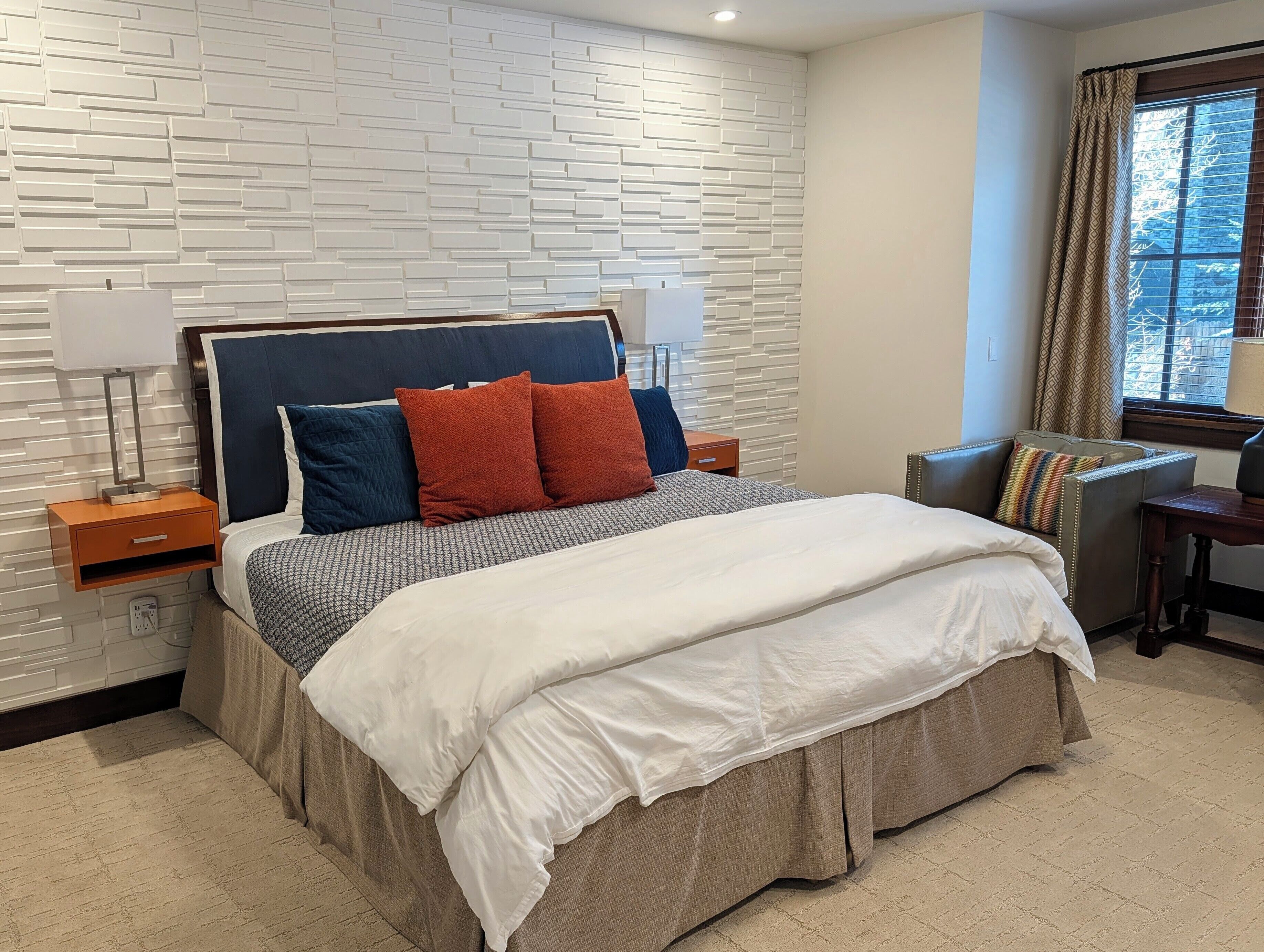 A modern hotel room with a bed, nightstands, a lamp, a window with curtains, and a view of trees.