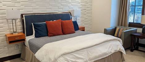 A modern hotel room with a bed, nightstands, a lamp, a window with curtains, and a view of trees.
