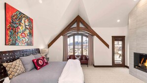 A bedroom with a large bed, a fireplace, a painting, and a view of mountains through a window. - Courcheval Chalet in Mt Village Core with use of 2 free ski passes during stay! (Mountain Village)