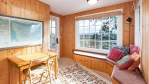 Une pièce chaleureuse aux murs en bois, avec une grande fenêtre offrant une belle vue, un bureau avec une chaise et un banc rembourré agrémenté de coussins.