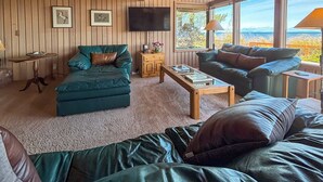 A cozy living room with a green sofa, a black sofa, and a wooden coffee table. There is a TV mounted on the wall and a window with a view of the outdoors. - Waterfront, Hot tub, Pets OK, Sequim Dungeness (Sequim)