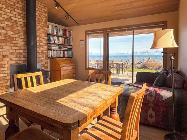 A cozy living area with a wooden table and chairs, a bookshelf, a fireplace, and a large window offering a view of the outdoors.