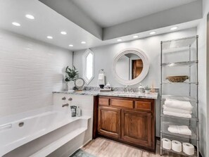 Ein modernes Badezimmer mit einer freistehenden Badewanne, Holzschränken, einer Marmorarbeitsplatte, einem runden Spiegel und einem Metallregal mit Handtüchern.