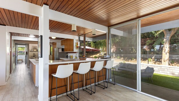 Una cocina moderna con techo de madera, una barra con sillas blancas y un amplio ventanal corredera que da a un patio trasero con césped y un árbol.