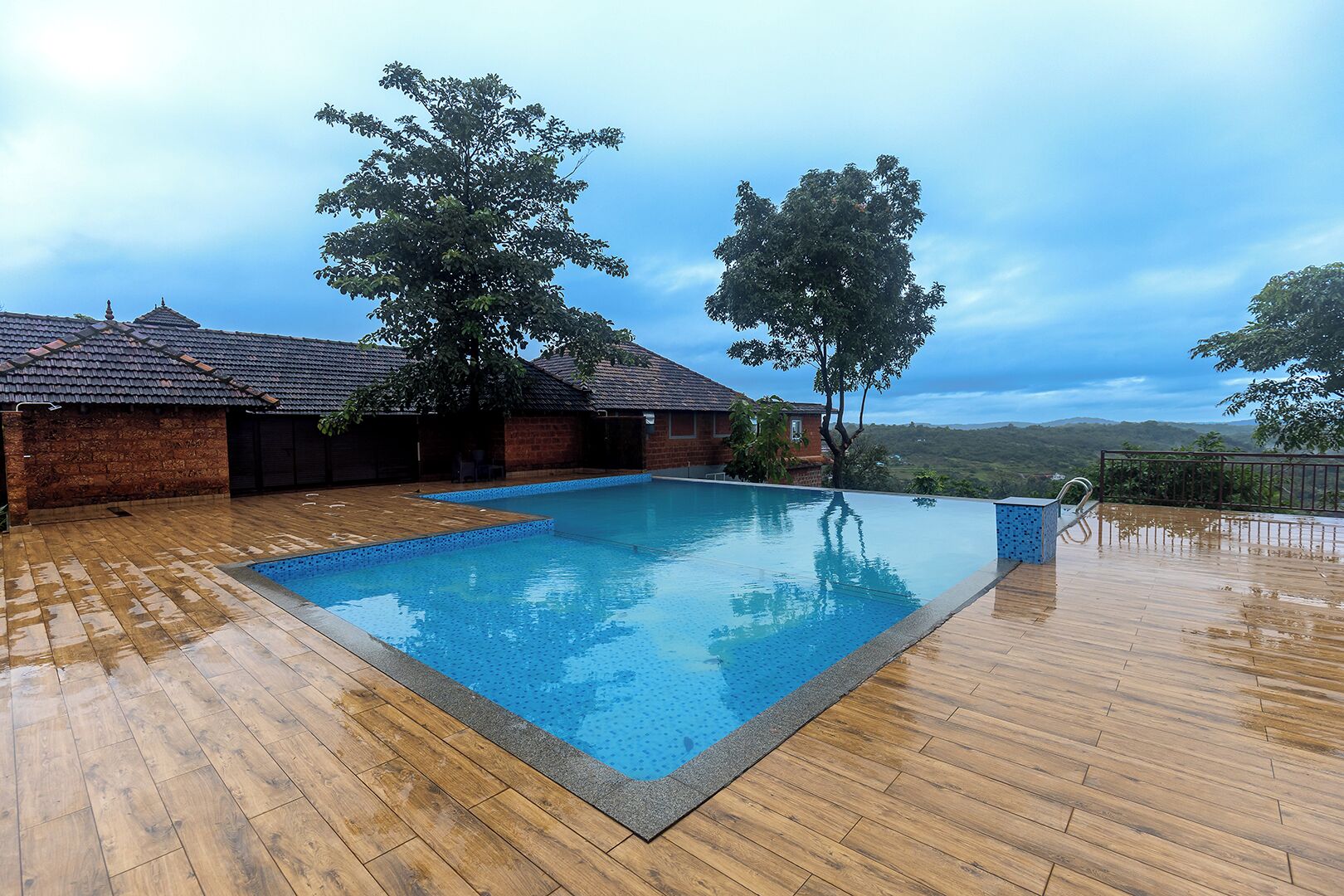 A rectangular swimming pool with a wooden deck surrounding it, a brick building with a tiled roof, and trees in the background.