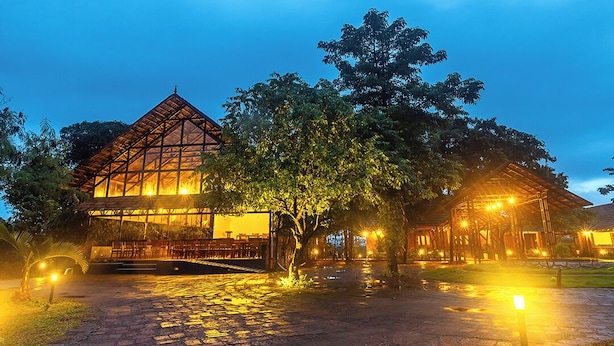 A building with a large glass facade and a wooden roof, surrounded by trees and illuminated by warm lights. - Sandal Mist Resort and spa  (Kasaragod)