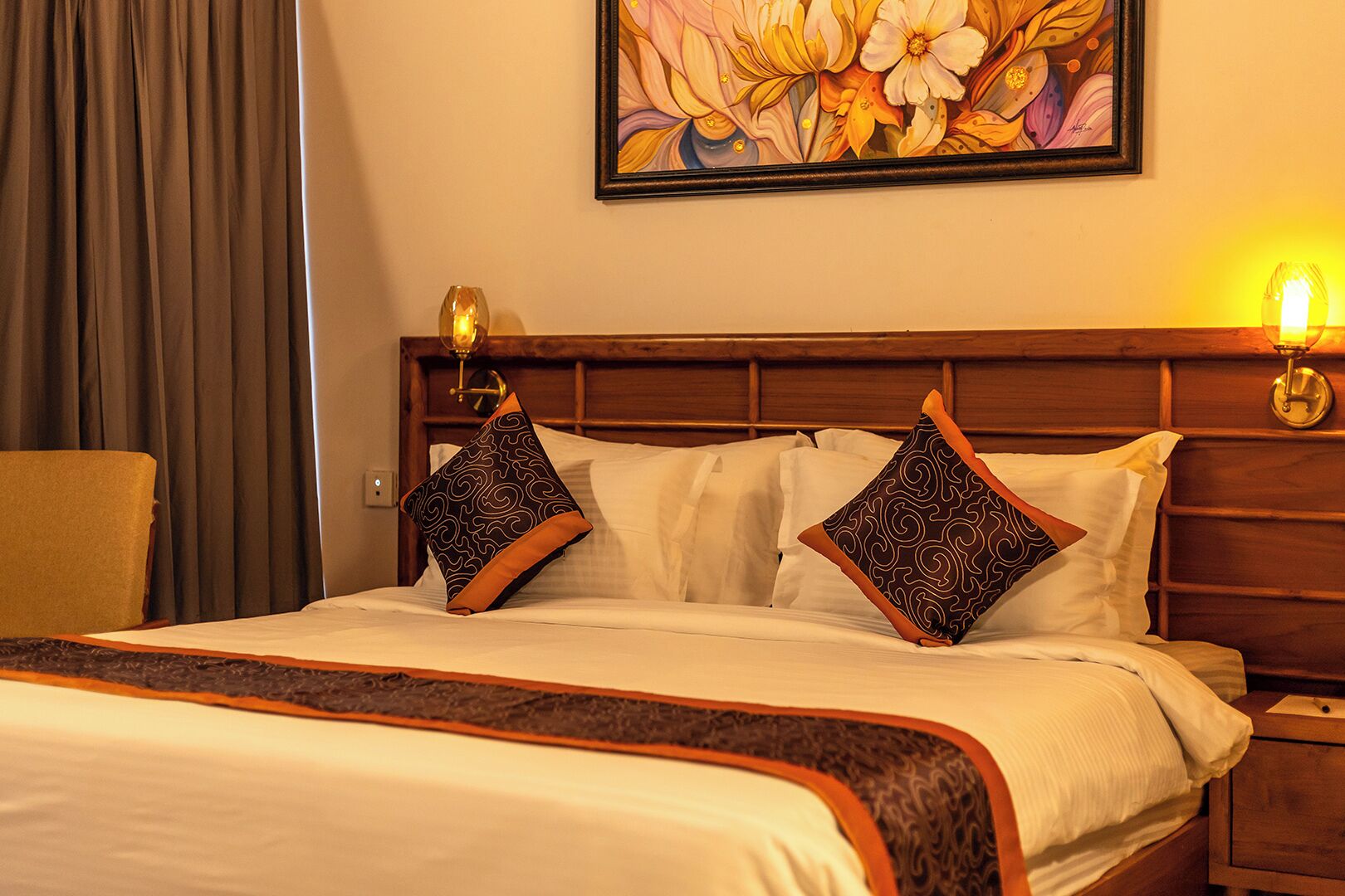 A hotel room with a wooden headboard, two patterned pillows, a white bedspread, a decorative border, a painting of flowers on the wall, and a lamp on the nightstand.