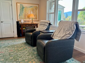 A room with two leather chairs, a wooden side table with a lamp, a framed map on the wall, and a view of greenery outside.