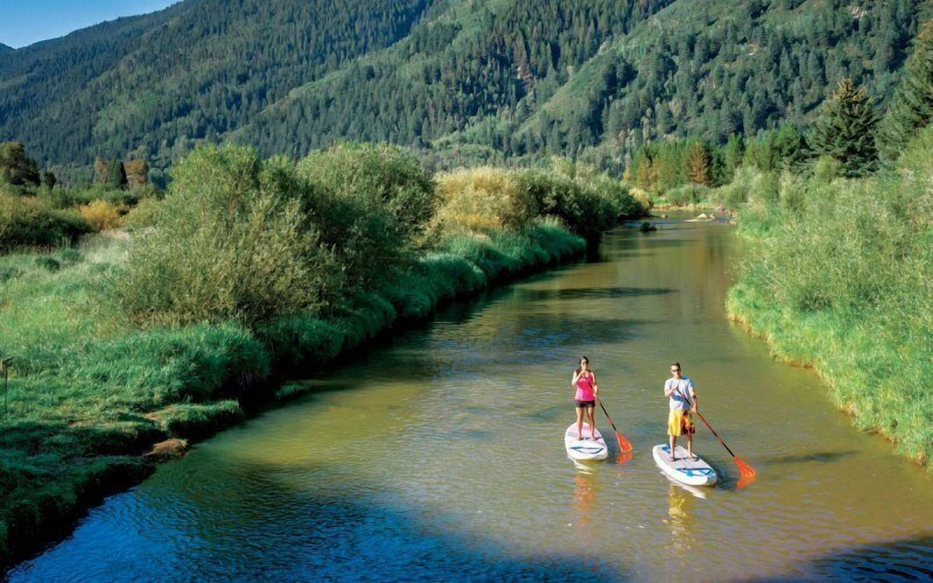 Duas pessoas praticando stand-up paddle em um rio calmo, cercadas por vegetação exuberante e montanhas.