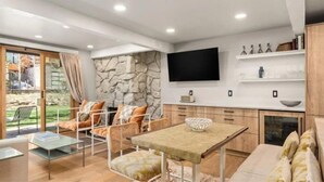 A modern living room with a stone wall, a flat-screen TV, a dining table, and a seating area with chairs. - Aspen 2 Bedroom Condo at the Base of Aspen Mountain, Next to Lift 1A (Aspen)