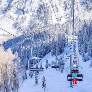 A ski lift with a single chair carrying a skier, surrounded by snow-covered trees and a mountainous backdrop. - Aspen 2 Bedroom Condo at the Base of Aspen Mountain, Next to Lift 1A (Aspen)