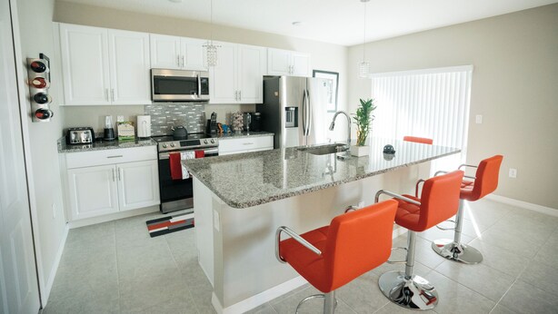 A modern kitchen with white cabinets, stainless steel appliances, and a granite countertop. There are three orange bar stools and a potted plant. - Welcome to your Serene Retreat.  (Palm Bay)