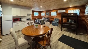 A cozy cabin interior with a wooden dining table, chairs, a kitchen area, a living room with a sofa and armchair, and a bunk bed. - Adorable log cabin on back country road with peace, quiet, all amenities of home (Kimbolton)