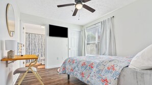 Un dormitorio con cama, un escritorio con silla, un ventilador de techo y un televisor en la pared.